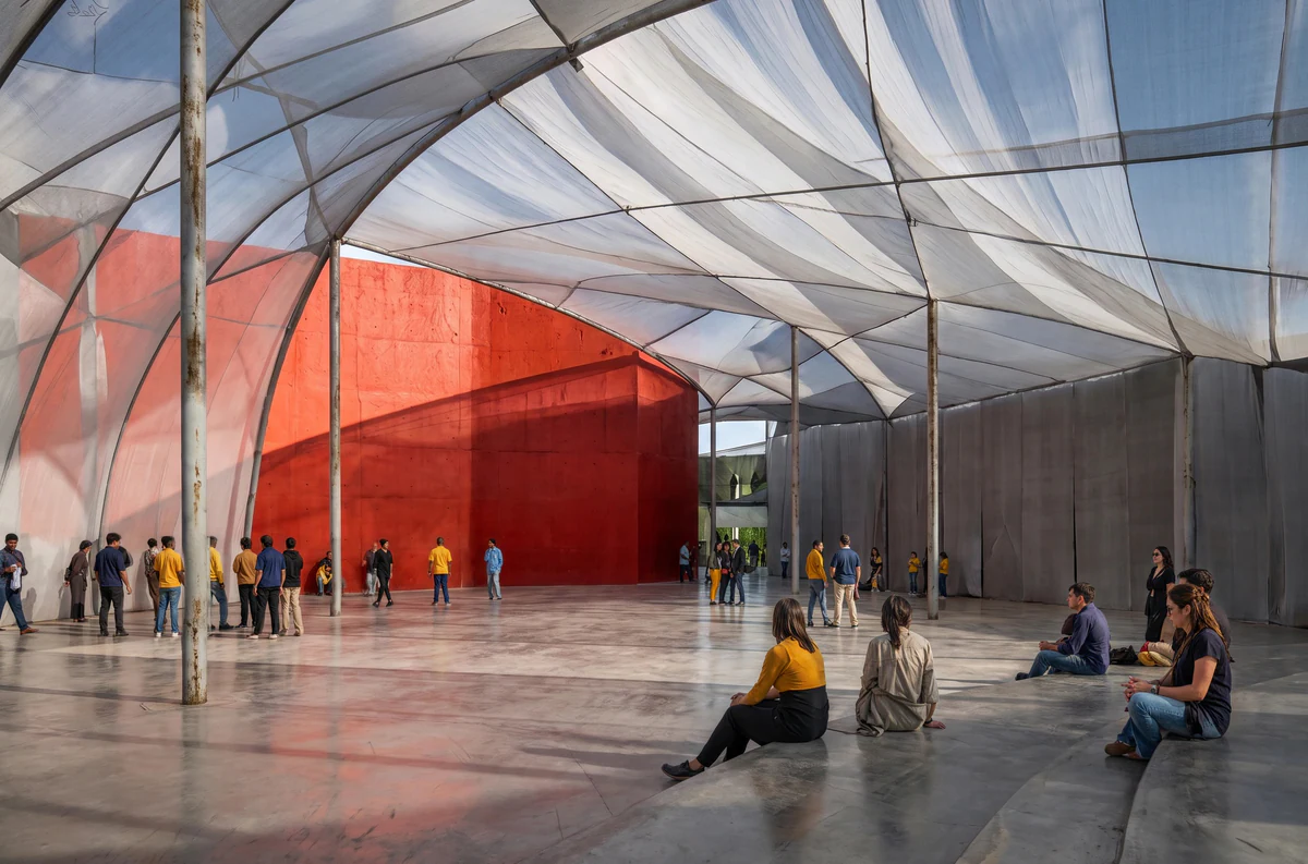 A crowd under a white tensile canopy on a polished concrete plaza, gathered around a red concrete volume.