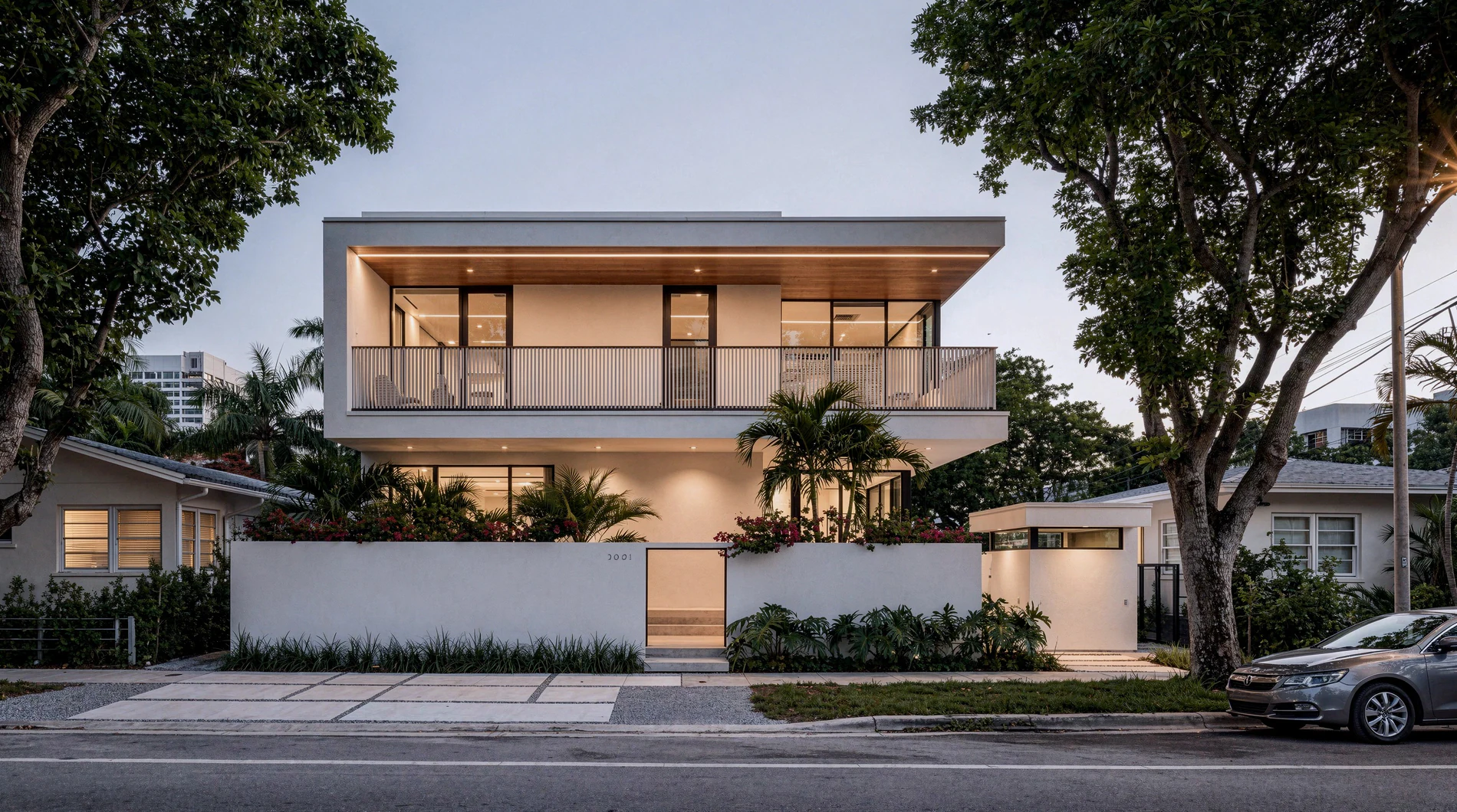 A lit two-storey house behind a white wall at dusk, palms and a street in front.