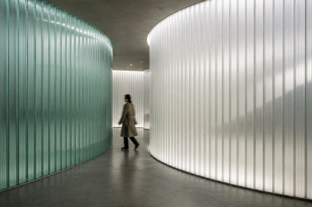A curved corridor walled in fluted green glass, one figure walking through it.